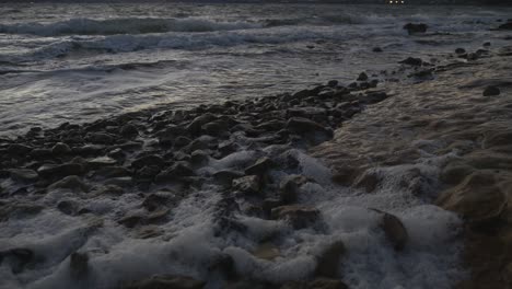 Cámara-Lenta-De-Olas-Rodando-Sobre-Rocas-En-La-Playa-Causando-Espuma-Oceánica-En-La-Noche-Ventosa-Del-Atardecer