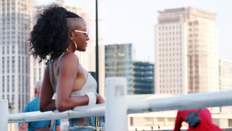 Fashionable-young-black-woman-leaning-on-a-bridge-in-the-city-looking-around,-back-view