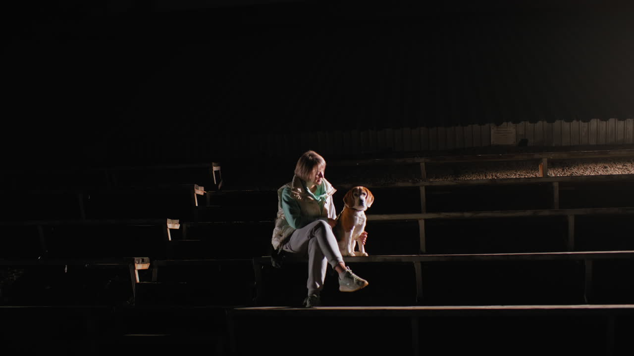 Female dog owner with legs crossed sits on football bench at night under spotlight sharing quiet moment with her pet beagle, both gazing calmly into distance surrounded by empty stadium stands