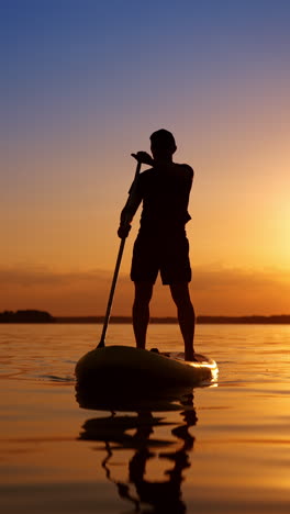 male silhouette holding an oar above his head. man oars by the river standing on sup board at sunset. low angle view. vertical video.