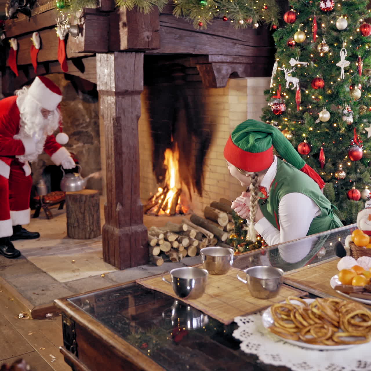 Santa Claus and an elve girl. Santa preparing tea. Santa Claus with boiling teapot, Curious girl wears elves clothes. Happy elf girl in fairy costume sitting near the table. Santa near Christmas fireplace.