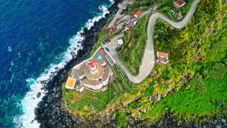 A-coastal-lighthouse-perched-on-a-cliff-with-winding-roads-and-lush-greenery,-aerial-view