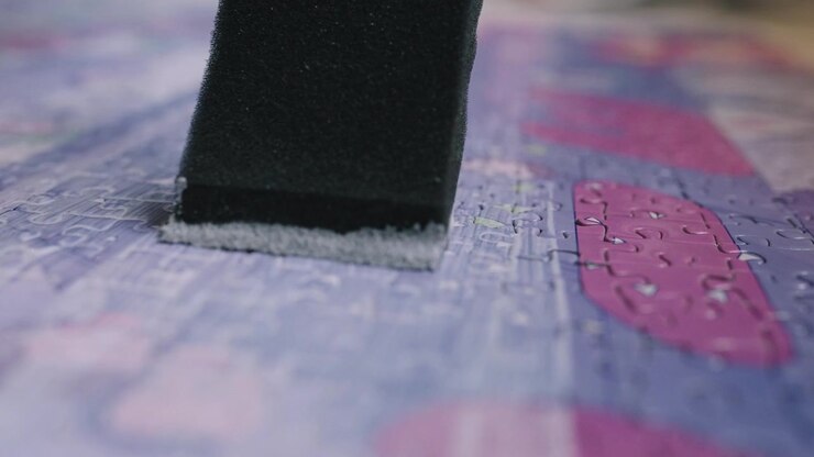 The camera slowly follows a hobbyist as they paint mod pudge puzzle glue onto a finished puzzle.
