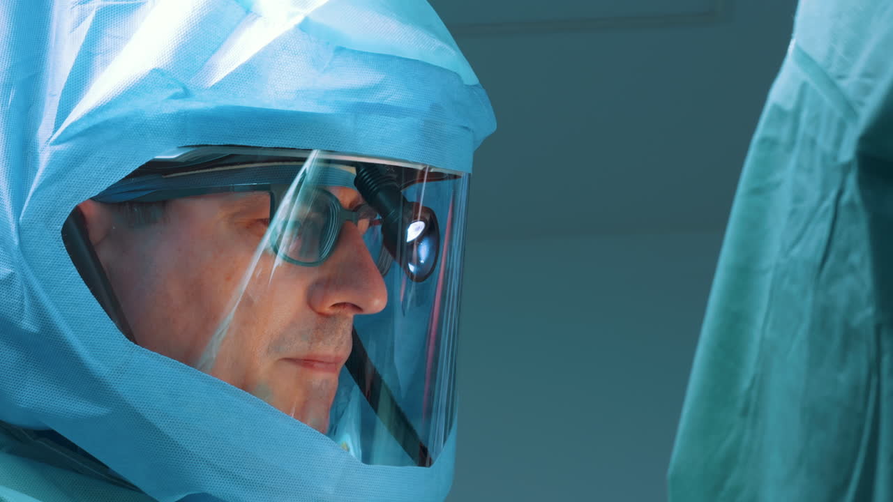 Close-up side profile of a male surgeon in a sterile helmet and head-mounted light. The professional's focused expression is visible through the face shield in the blue-lit environment