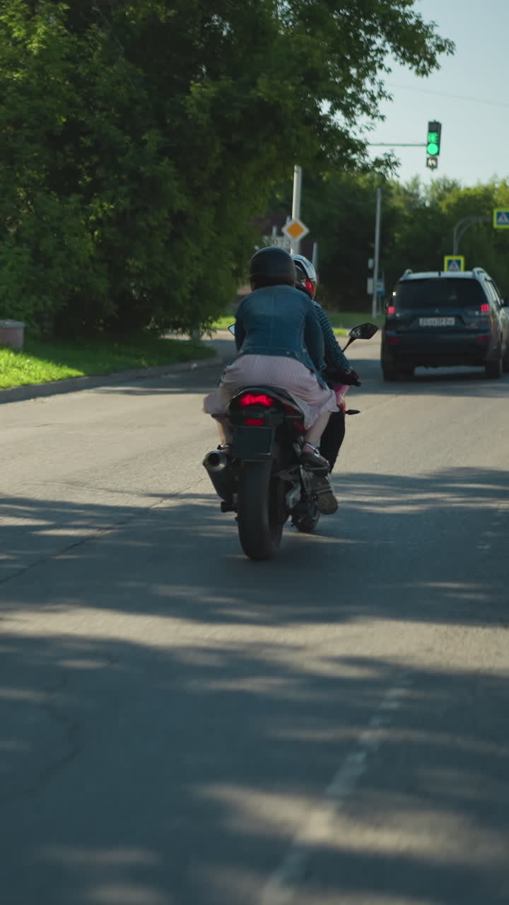 vista trasera de dos amigos montando una bicicleta eléctrica, siguiendo dos coches con un indicador de semáforo verde delante, un tranvía también es visible en el lado mientras viajan a lo largo de una carretera urbana sombreada en un día soleado