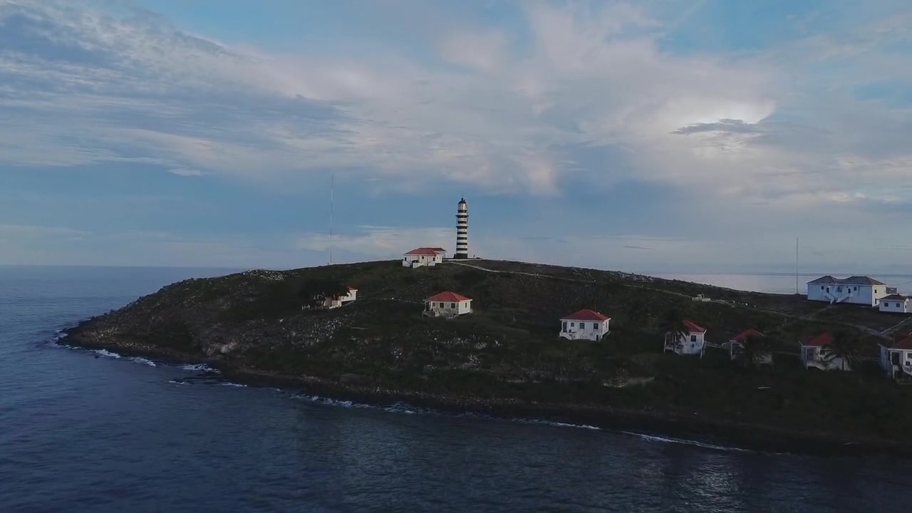 Farol de Abrolhos sull'isola di Santa Bárbara nel Parco Nazionale Marino di Abrolhos, Caravelas, Bahia, Brasile, con faro, alloggi del personale e costa rocciosa sotto un cielo nuvoloso, ripresa con drone in volo in avanti.