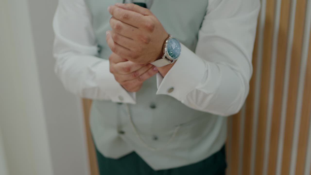 Groom in white shirt and light gray vest adjusting silver mesh watch before wedding
