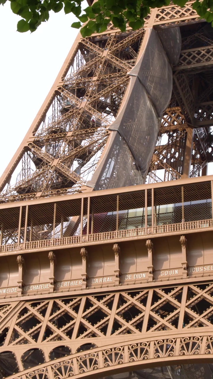 Inquadratura dal basso della struttura della Torre Eiffel con foglie verdi che incorniciano il bordo superiore.