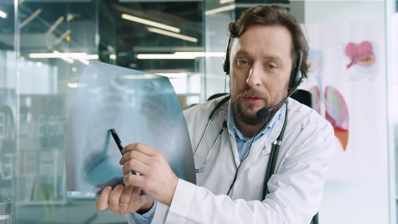 Close-up view of senior male doctor with headphones sitting at desk speaking at camera and explaining coronavirus lung disease in hospital office