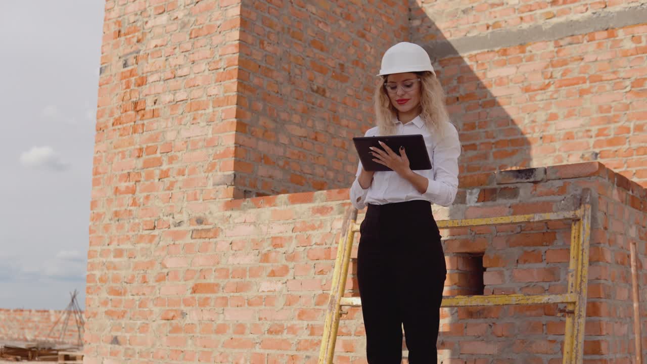 arquitecta femenina en ropa de negocios y un casco blanco se encuentra en una casa de nueva construcción con paredes no tratadas y trabaja en una tableta. tecnologías modernas en las profesiones más antiguas