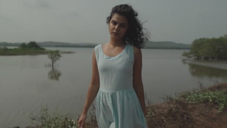 slow motion panning shot of a young and elegant lady in light blue dress with black hair standing in front of a lake with plants and looking into the camera with an appealing look