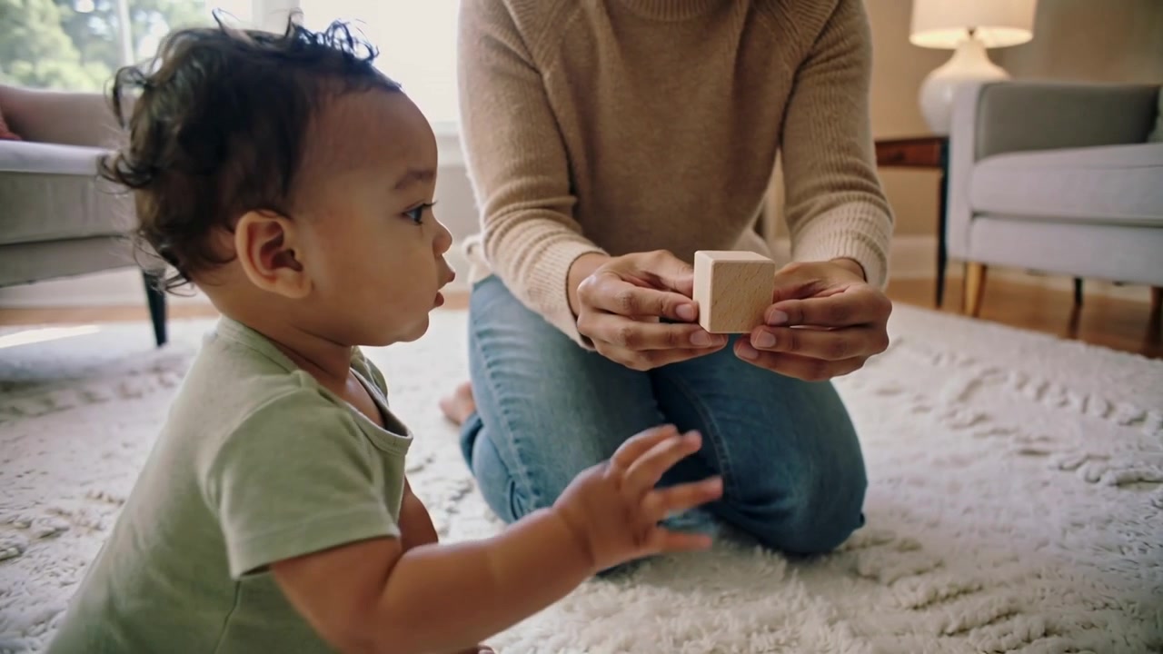 Kneeling parent holding wood cube to infant in mint onesie on living room rug, encouraging grasping
