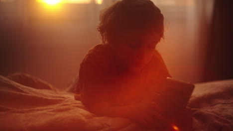 child lying on bed, looking at smartphone at sunset