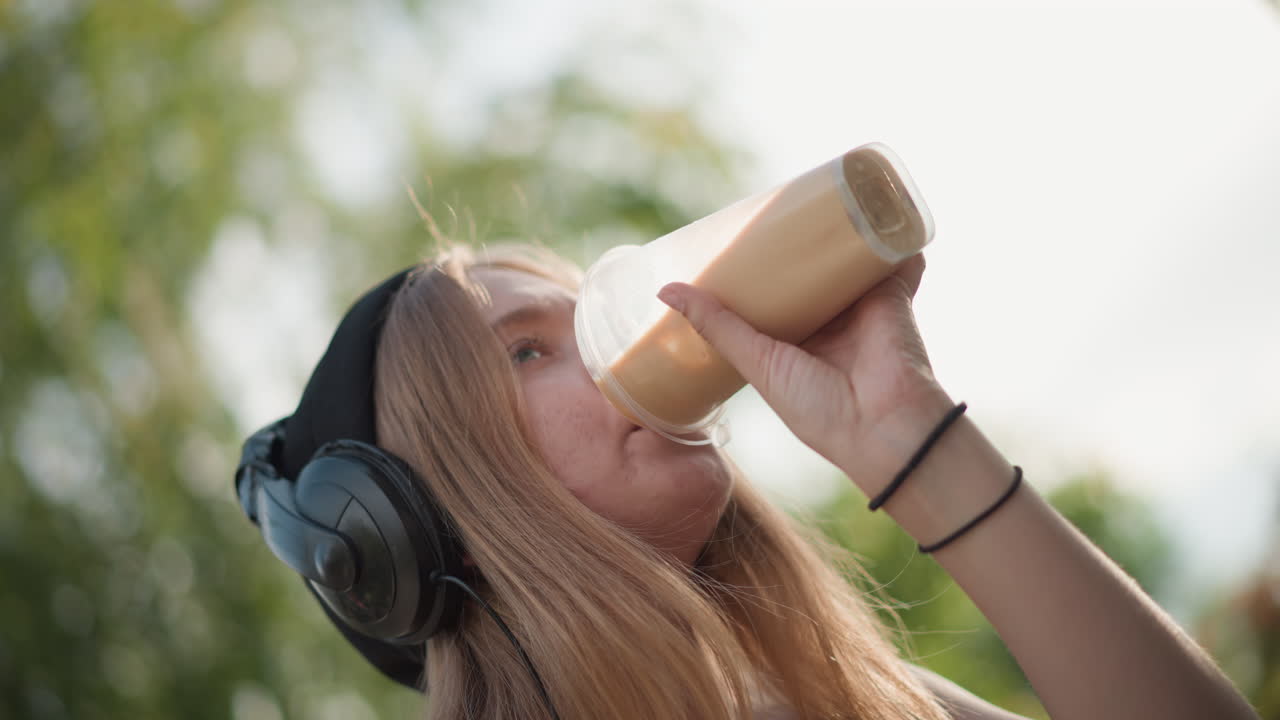 Giovane donna beve da una bottiglia all'aperto, sorseggiando un frullato mentre sistema le cuffie, battendo il piede a ritmo di musica, sorridendo verso il cielo, calda luce del sole tra gli alberi, scena di un tavolo da picnic, primo piano