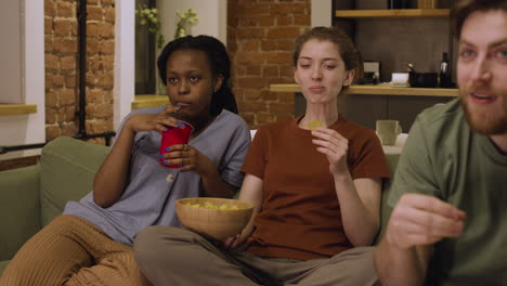 tres compañeros de cuarto están viendo una película mientras comen papas fritas sentados en el sofá en casa