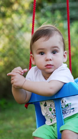 sweet caucasian baby boy with bare feet sits in a swing. cute child having good time outdoors in summer. blurred nature backdrop. vertical video