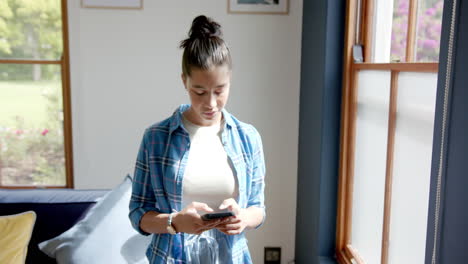 una adolescente biracial feliz de pie en una habitación soleada usando un teléfono inteligente, espacio de copia, cámara lenta