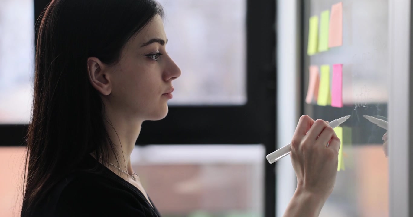 Donna bruna concentrata scrive appunti su una parete di vetro in ufficio