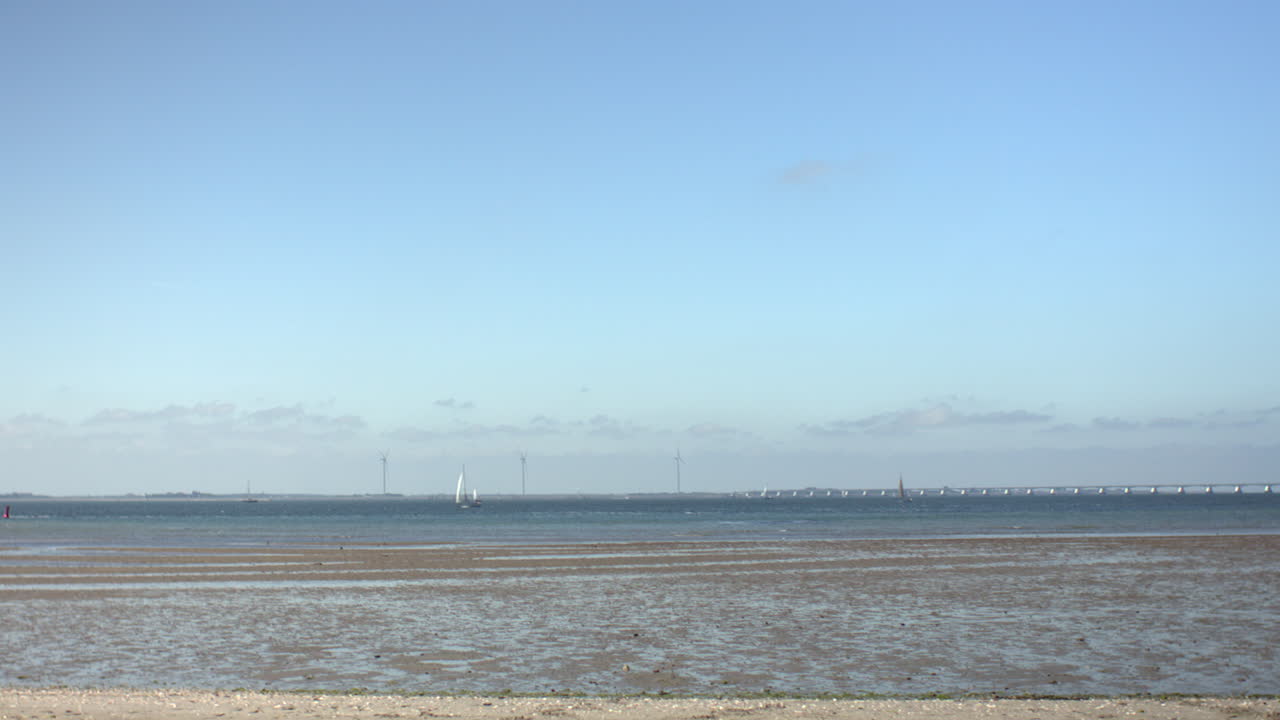 Wide view of the Dutch coast with shallow tidal water, a distant sailboat, modern wind turbines, and a long bridge across the horizon. Peaceful scene from a coastal viewpoint