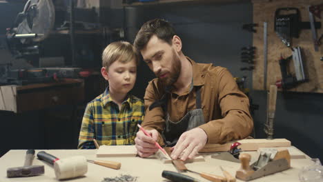 Joven-Carpintero-Caucásico-Enseñando-A-Trabajar-Con-Madera-A-Su-Pequeño-Hijo-En-El-Taller