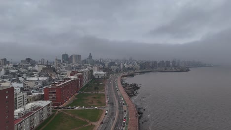static aerial shot of fog and low clouds moving over montevideo’s rambla sur on a moody gray afternoon. city skyline fades as weather shifts over the coastline