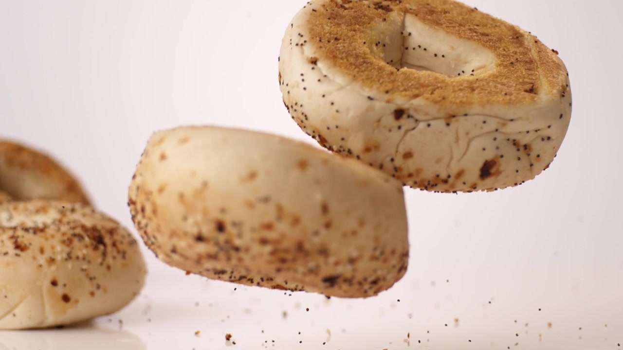Pile of everything-seeded savory bagels tumbling and bouncing on bright white backdrop in slow motion. office catering posts, weekend brunch reels, deli case branding.