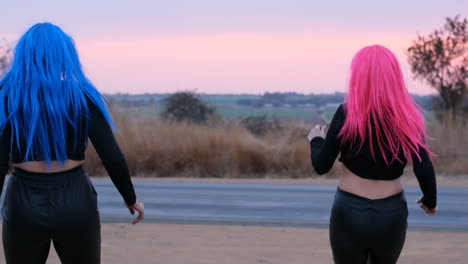 dos mujeres con pelucas rosas y azules corriendo hacia una calle transitada al atardecer