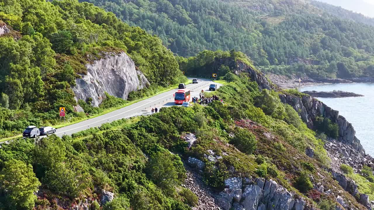 Aerial view of cars and bus traveling on winding coastal road, lush hills, bright daylight