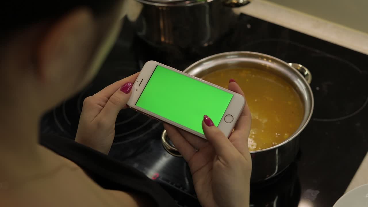 mujer está sosteniendo un teléfono inteligente con una pantalla verde, y cocinando sopa