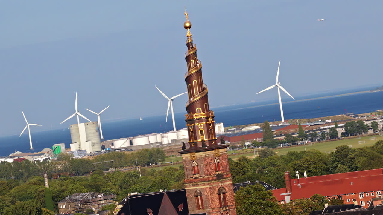 Aerial drone view of the Church of Our Saviour with its golden spiral tower, set against a backdrop of wind turbines and the sea in Copenhagen, Denmark