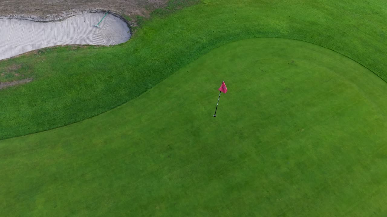 Drone flies low from a bunker toward the golf flag, then rises high above the green. The camera tilts down into a top-down view and rotates, ending directly over the green