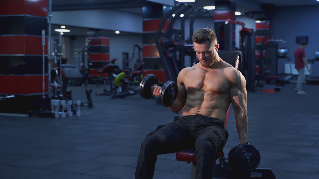 Bodybuilder professionista che si allena in palestra. Atleta muscoloso a torso nudo che allena i suoi muscoli con i manubri mentre è seduto su una sedia in un centro sportivo