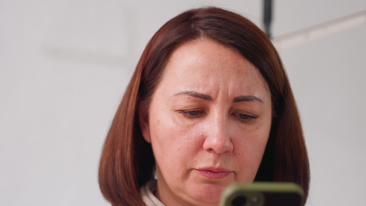 Close up of elegant woman intently speaking while focused on smartphone screen in bright indoor workspace, brows slightly furrowed, displaying concentration and serious mood during mobile interaction