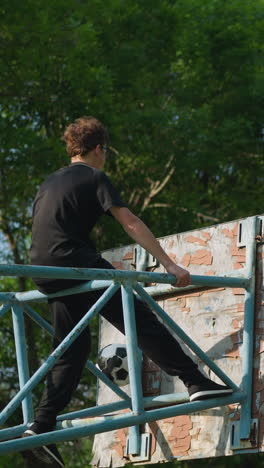 una vista trasera de un niño sentado en una estructura de hierro azul al aire libre, enfocado mientras agarra cada lado del marco de hierro, una pelota de fútbol desgastada está atascada en la estructura