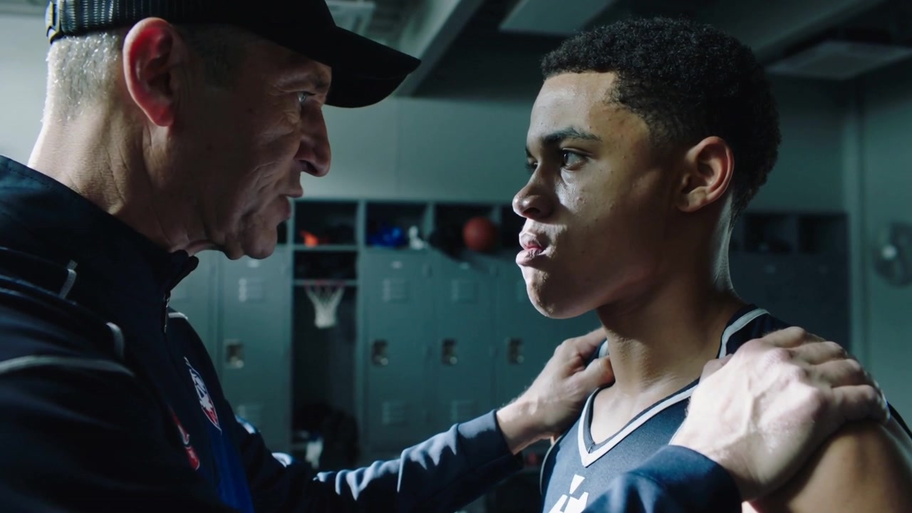 Stepping coach gripping teen shoulder in jersey seeking focus speaking in locker room by basketball