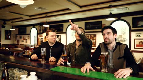 cheerful young men are watching football match, cheering and drinking beer while sitting in modern sports bar. mates are emotional and happy.