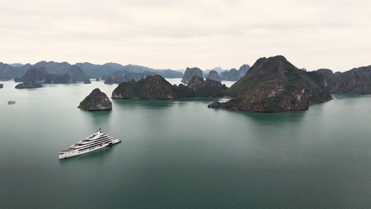 White luxury cruise ship glides through emerald waters among towering limestone karsts of Ha Long Bay, Quang Ninh Province, Vietnam, during cool dry season, captured by aerial drone shot.