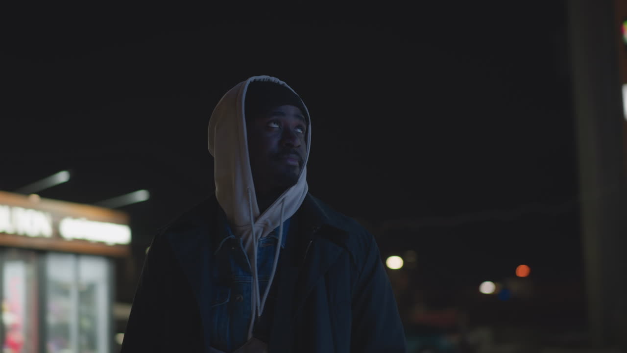 Close-up of man standing outdoors at night, wearing hoodie and jacket, illuminated by modern city lights in background, urban street setting with glowing signage