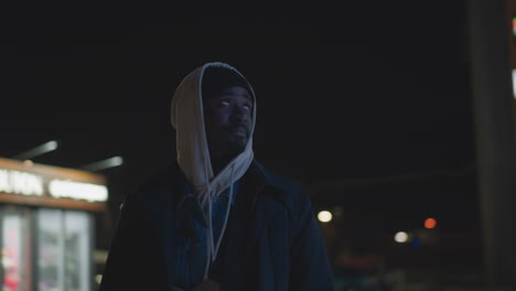 close-up of man standing outdoors at night, wearing hoodie and jacket, illuminated by modern city lights in background, urban street setting with glowing signage