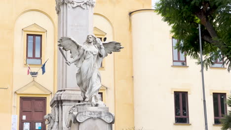 una estatua de ángel de mármol con alas extendidas se encuentra frente a un edificio histórico, que simboliza la paz o el recuerdo, con banderas italianas