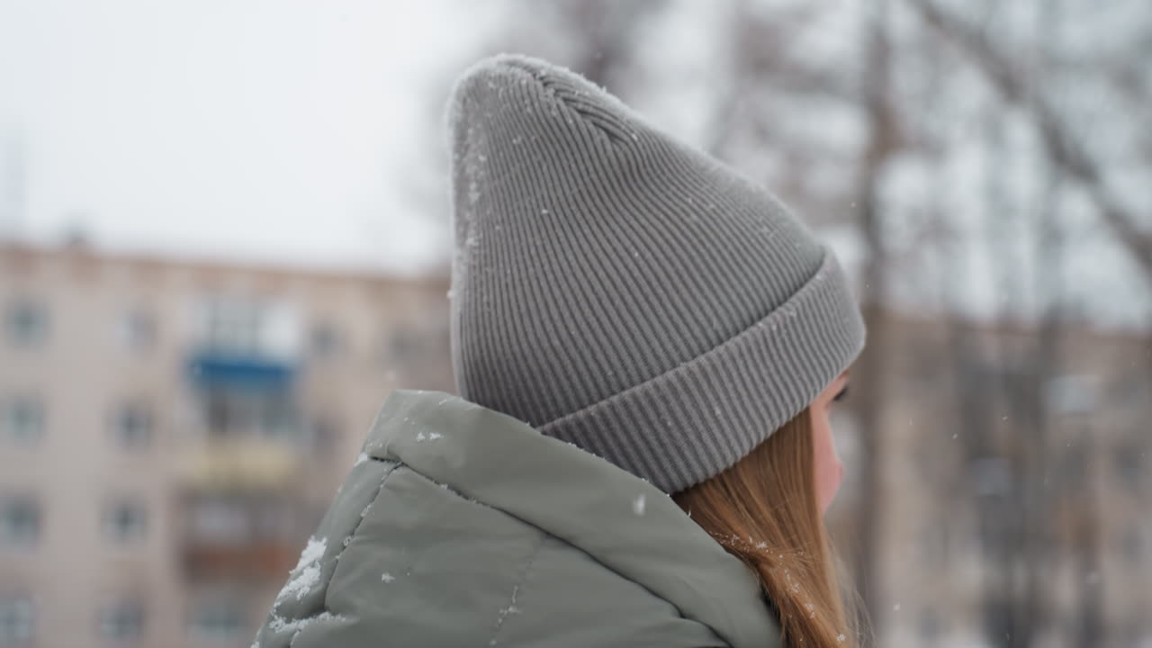 Dog owner in gray beanie and green winter jacket seen in profile smiling gently during snowy outdoor moment, snowflakes falling around, background blurred with trees and buildings in cold urban winter setting