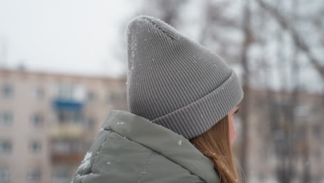 dog owner in gray beanie and green winter jacket seen in profile smiling gently during snowy outdoor moment, snowflakes falling around, background blurred with trees and buildings in cold urban winter setting