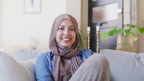 Videoporträt-Einer-Gemischtrassigen-Frau-In-Braunem-Hijab,-Die-Zu-Hause-Im-Wohnzimmer-In-Die-Kamera-Lacht