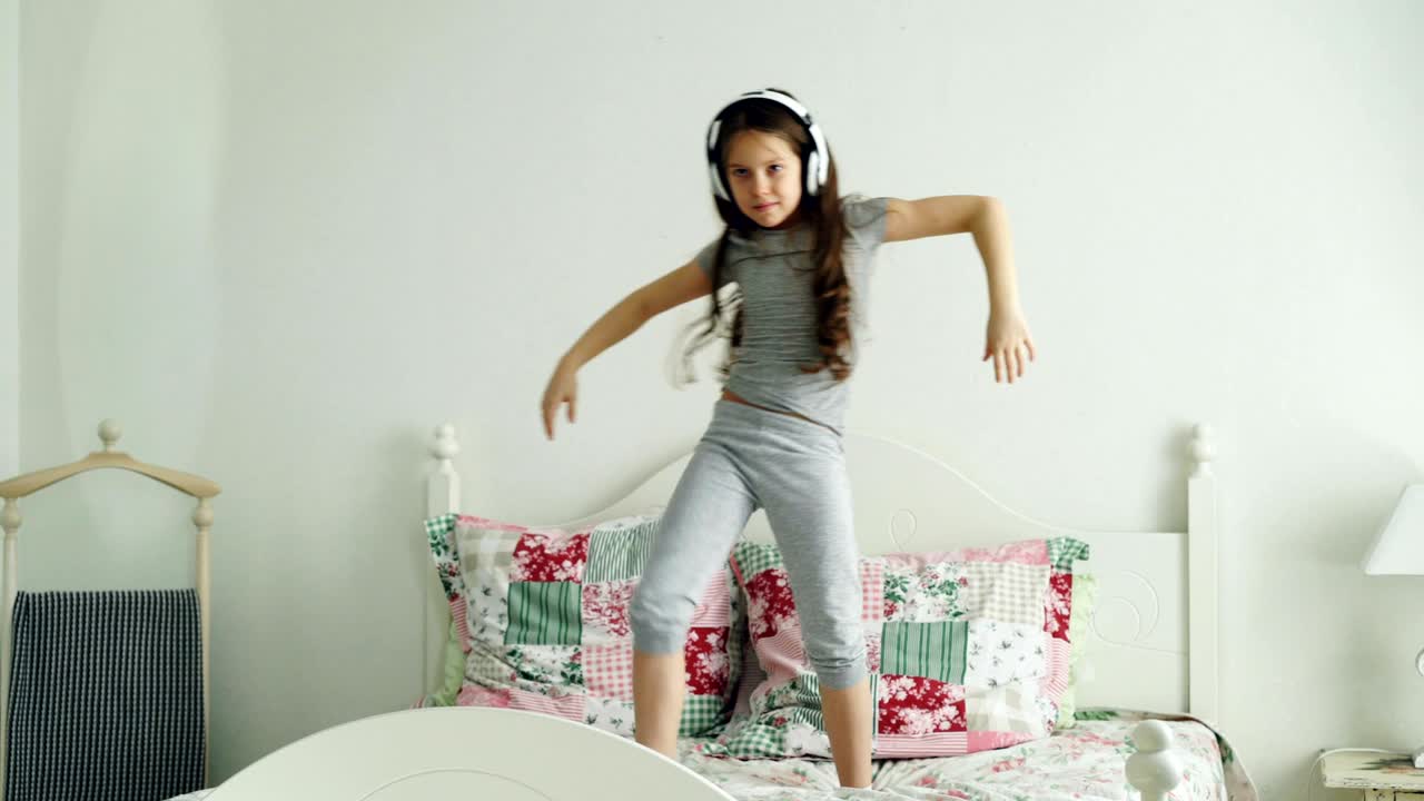 Simpatica bambina carina con cuffie wireless che balla e si diverte in una mattina di vacanza, saltando sul letto a casa in una camera da letto accogliente e luminosa.