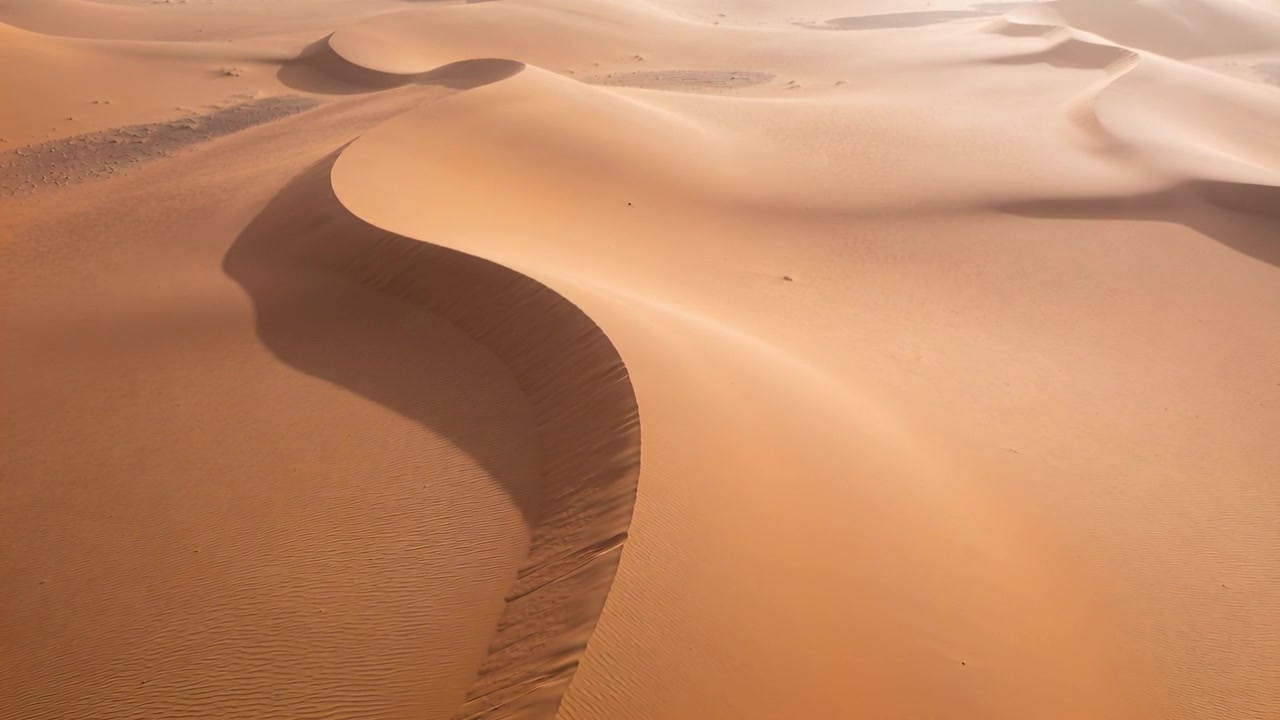 Drone che si lancia iniziando un lento passaggio in avanti sopra il deserto, rivelando increspature a forma di S sulla cresta delle dune.