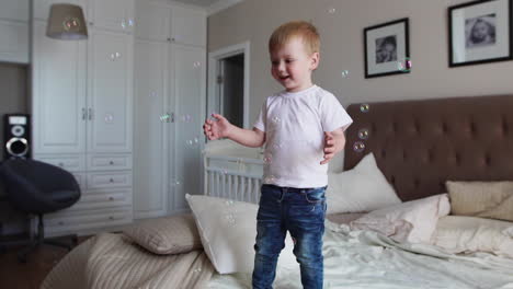 a boy in a white t-shirt and blue jeans catches soap bubbles standing on the bed in the parent bedroom