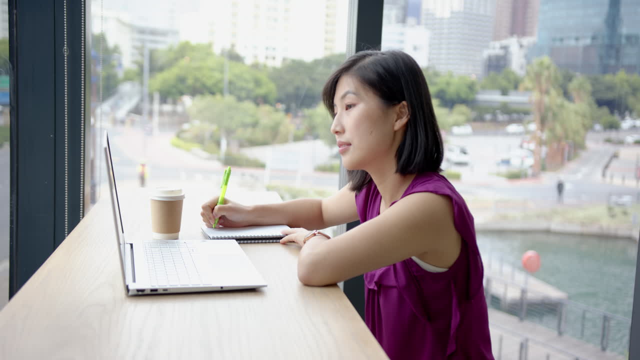 Writing in notebook, woman working on laptop with coffee cup in office