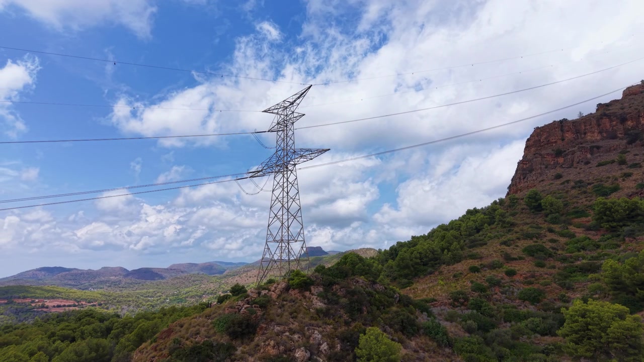 High voltage electricity pylon standing on a green hillside in a mountainous landscape under a blue sky with white clouds, red rocky cliffs visible to the right