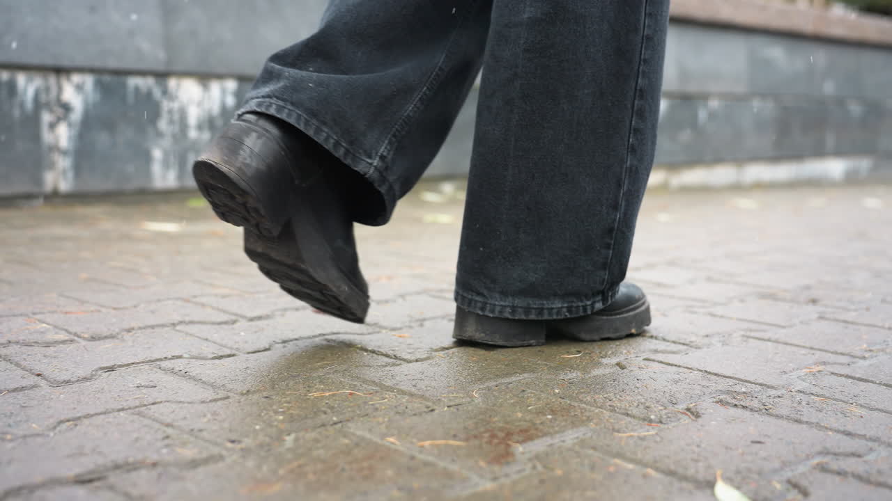 Close up of human leg wearing black trousers and black boots walking slowly on outdoor paved path during calm overcast day, capturing foot movement and subtle step motion