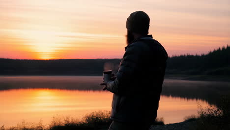 man enjoys warm drink by a scenic lake at sunset or sunrise